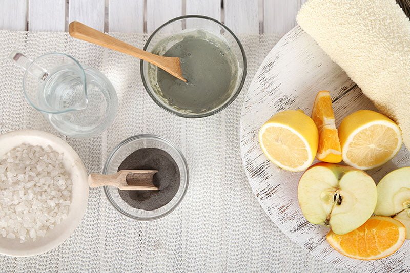 Top view of a spa setting with natural ingredients including lemons, apples, oranges, sea salt, clay mask, charcoal powder, and water. towel and wooden spoons add to the clean, refreshing ambiance.